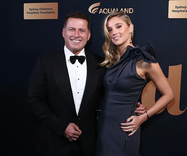 Elegant couple posing at a formal event, with a dark backdrop featuring sponsor logos.