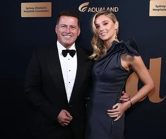 Elegant couple posing at a formal event, with a dark backdrop featuring sponsor logos.