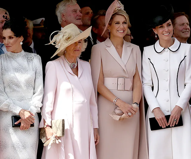 Women in elegant dresses and hats smiling at a formal event, standing in a row.