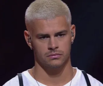 A person with short blond hair, wearing a white shirt and earpiece, looks serious on stage during a performance.