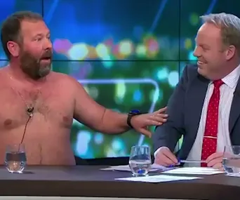 Shirtless man and suited man laughing on a TV talk show set with colorful background lights.