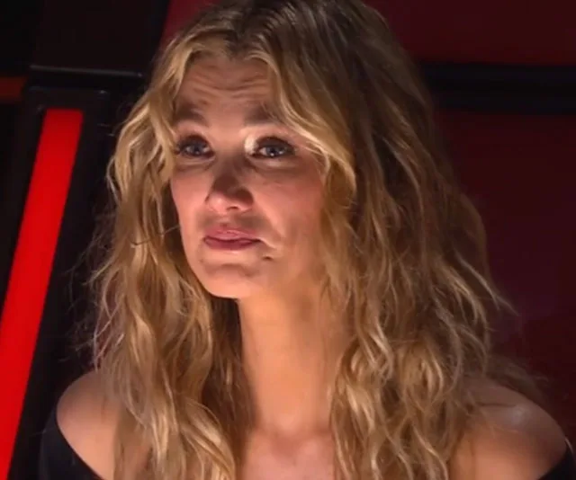A person with wavy blonde hair and a solemn expression, seated in a dimly lit room, possibly on a television show set.
