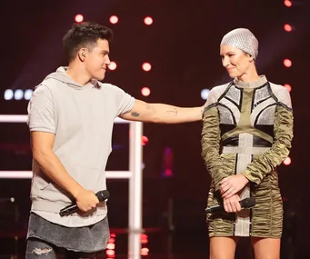 Two contestants on stage holding microphones, one with short hair and a gray hoodie, the other in a shiny, fitted dress.