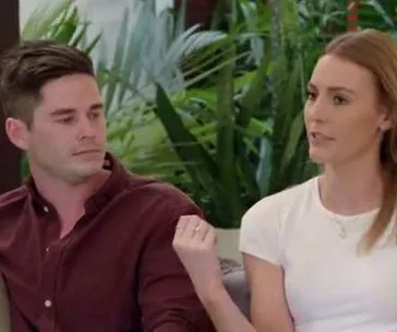 Two people seated indoors, a woman in white gesturing while a man in a maroon shirt listens, with plants in the background.
