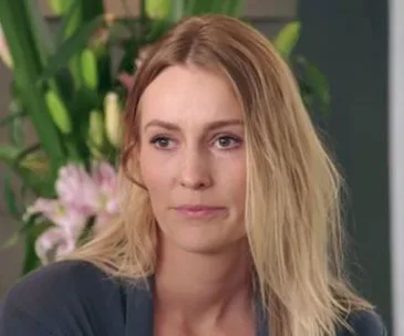 Blonde woman with long hair in front of green plants, wearing a gray top, looking to the side with a neutral expression.