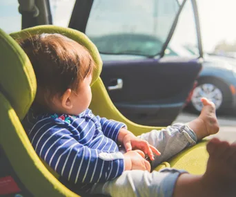 Baby in striped shirt sitting in green car seat, looking out the open car door toward sunlight.