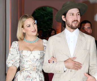 Kate Hudson in a floral dress with necklace, with a man in a light suit and hat, at an outdoor event.