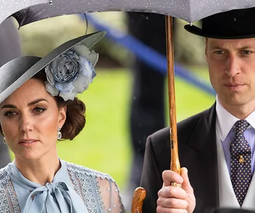 Royal couple under an umbrella, dressed formally, with the woman wearing a hat adorned with a large flower.