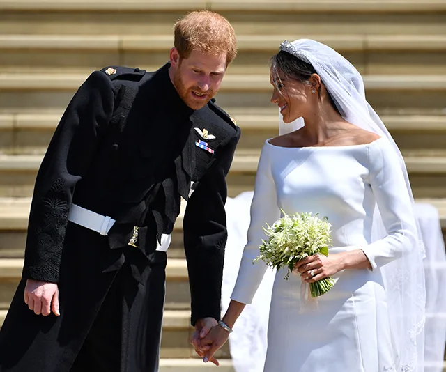 Prince Harry and Meghan Markle smile and hold hands on their wedding day, Meghan in a white gown.