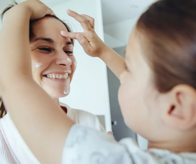 Mother and child smiling as the child applies cream to mother's face in a playful skincare routine.