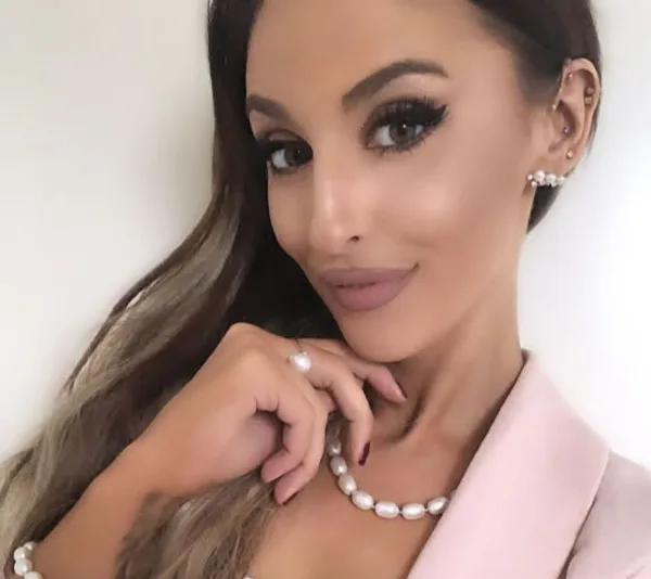 Lizzie in a pink outfit, wearing pearl earrings and necklace, posing with a soft smile against a plain background.