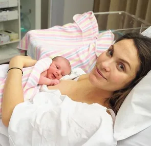 A mother in a hospital bed smiles at the camera while holding her newborn wrapped in a pink striped blanket.
