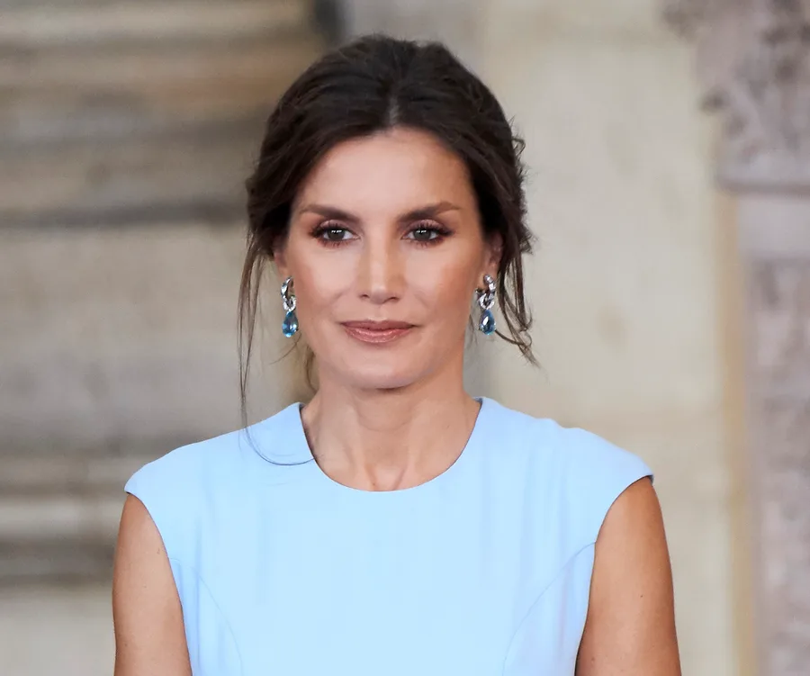 Woman in a light blue dress and blue earrings, standing indoors with an elegant hairstyle.