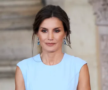 Woman in a light blue dress and blue earrings, standing indoors with an elegant hairstyle.