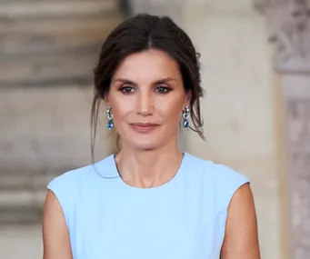 Woman in a light blue dress and blue earrings, standing indoors with an elegant hairstyle.