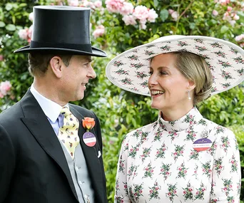 Couple in formal attire and floral hats smiling at each other amidst a backdrop of pink flowers.