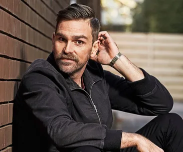 A person sitting against a brick wall, wearing a dark jacket, looking off-camera thoughtfully.