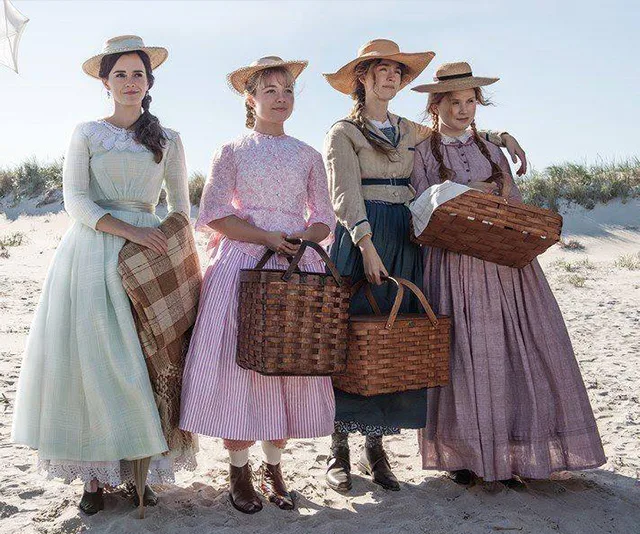 Four women in period dresses and straw hats stand on a beach, each holding a wicker basket.