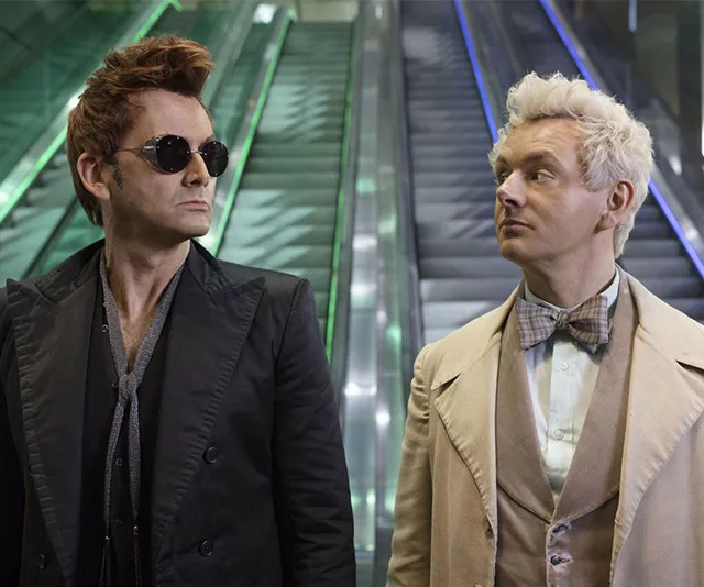 Two men in contrasting outfits, one in black with sunglasses and the other in light colors with a bow tie, in front of escalators.