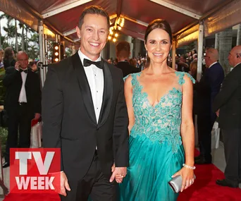 A man in a tuxedo and a woman in a teal lace gown smile on the red carpet at a formal event.