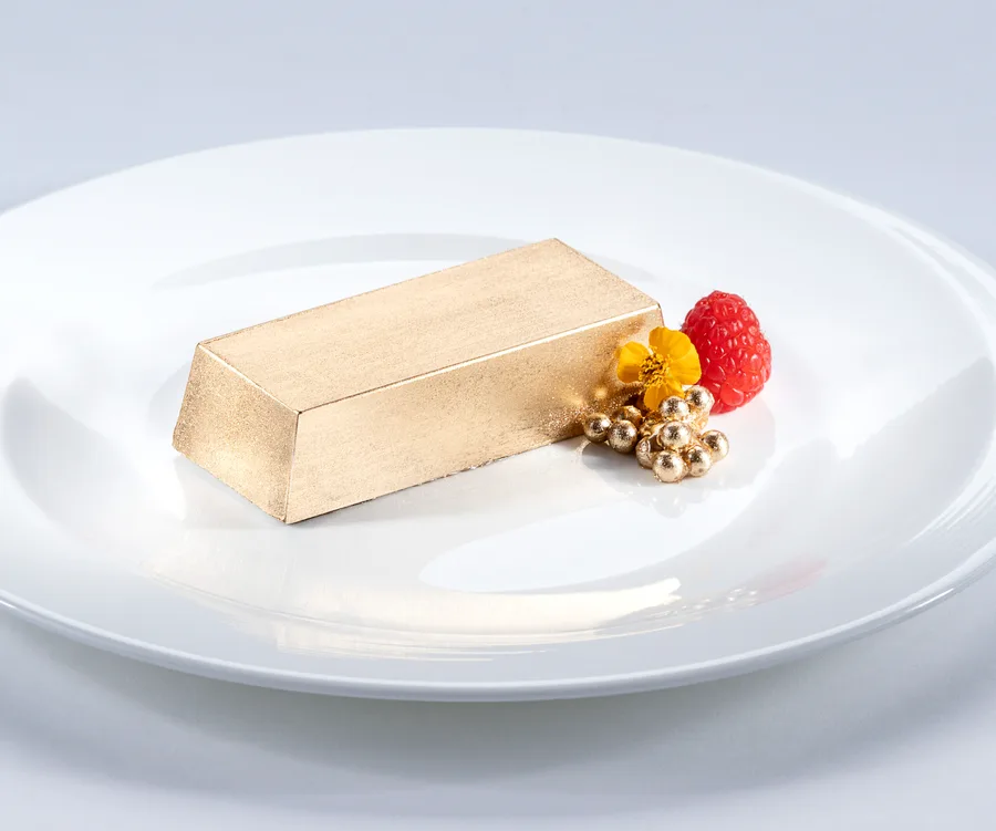 Gold dessert bar on white plate with raspberry and gold pearls, decorated with a yellow flower.