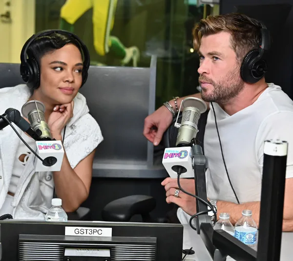 Chris Hemsworth and co-star at a radio station wearing headphones, speaking into microphones.