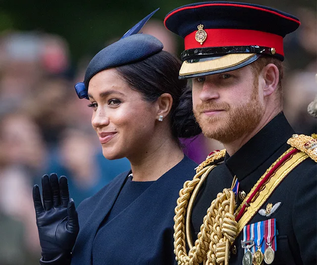 A man and woman in formal attire at a public event. The woman waves gently and the man wears a military uniform and hat.