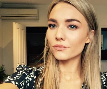 A woman with long blonde hair takes a selfie indoors, wearing a patterned top.