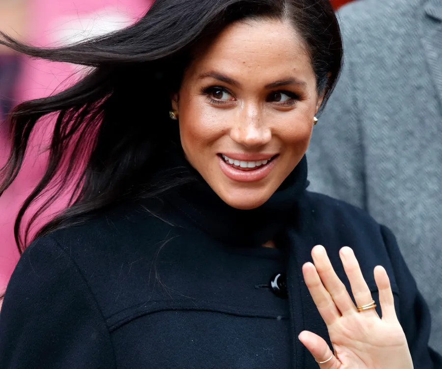 Meghan Markle smiling and waving, wearing a black coat, with hair blown by the wind.