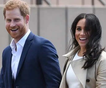 Prince Harry and Meghan Markle smiling, dressed in formal attire outdoors.