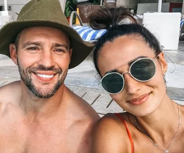 A couple smiling at the camera; the man wears a hat, the woman wears sunglasses.