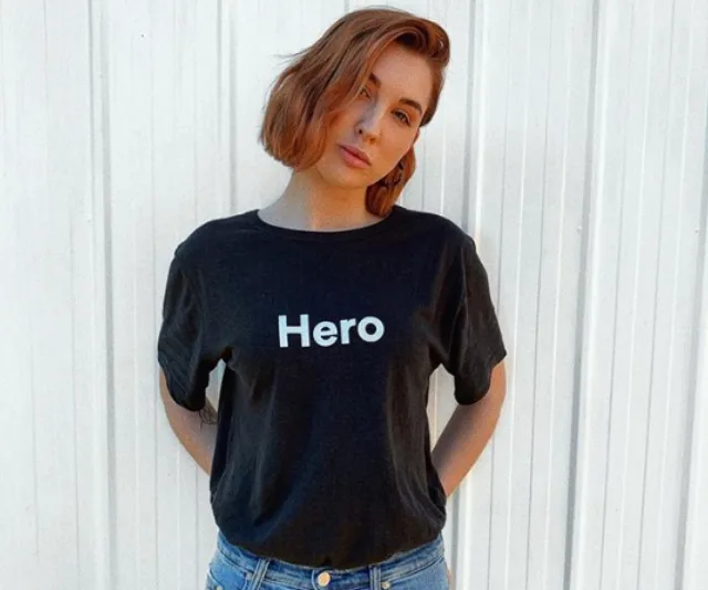 Woman with short hair wearing a "Hero" t-shirt, standing against a white wall.