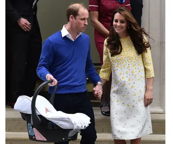 Couple walking out of a building, holding hands, with one carrying a baby in a car seat.