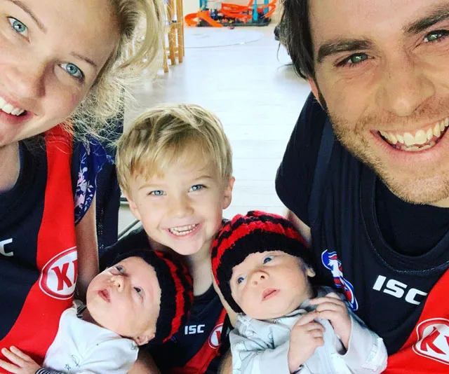 Family posing together, smiling; two parents, an older child, and two infants wearing knit hats.