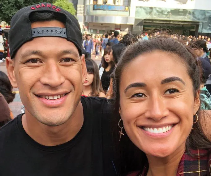 A couple smiling outdoors, with a crowd in the background on a sunny day.