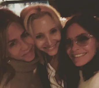 Three women smiling together in a close-up group selfie, warmly lit setting.