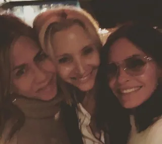 Three women smiling together in a close-up group selfie, warmly lit setting.