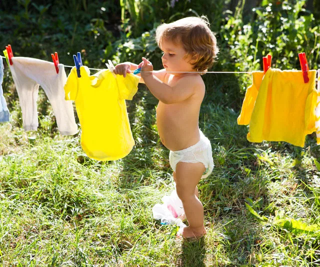 Toddler in diaper hanging a yellow shirt on a clothesline outdoors.