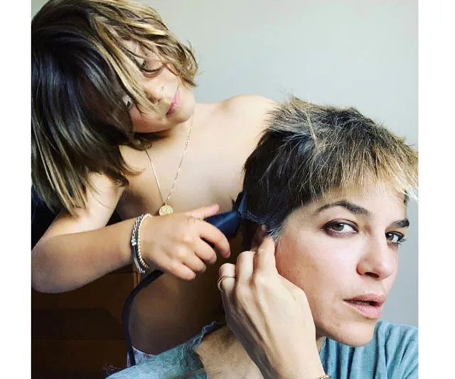 A young child shaves a woman's head with clippers, both looking engaged in the haircut process.