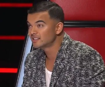 A person with short dark hair in a stylish grey jacket sits in a red chair, seemingly on a TV show set.