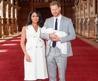 A couple stands in a formal hall holding a baby, both smiling at the camera.