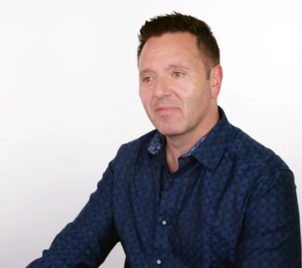 A man in a blue patterned shirt sits against a plain white background.