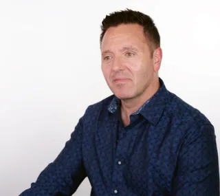 A man in a blue patterned shirt sits against a plain white background.