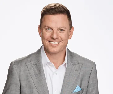 A man in a gray suit smiling at the camera with a white background.