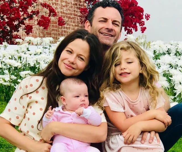 A family of four sits together in a garden with flowers; parents and two children are smiling.