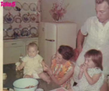 A family in a vintage kitchen, with a woman holding a baby on a counter, a man standing, and a young girl eating a snack.