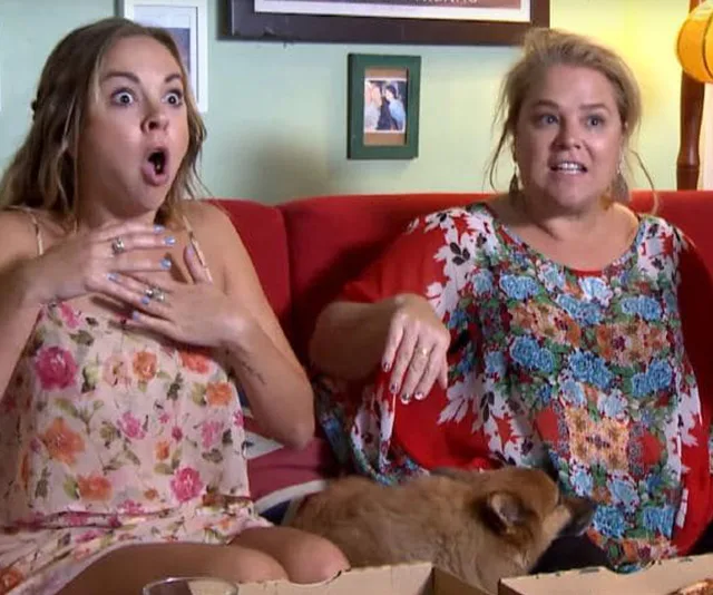 Two women on a couch, expressing shock and surprise, with a dog lying on their lap.