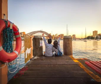 Couple in matching outfits sitting on a dock facing Sydney Harbour Bridge at sunset, with sailboats in view.
