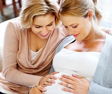 Two women smiling, one is pregnant with her friend touching her belly, showing affection and joy.