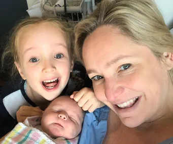 A woman smiles with her newborn baby and young child, who appears excited, all lying close together.